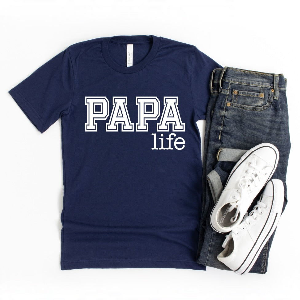 The Papa Life • Navy Tee, a Bella Canvas unisex essential shirt with bold white PAPA life print, is styled on a white background alongside folded blue jeans and white sneakers.