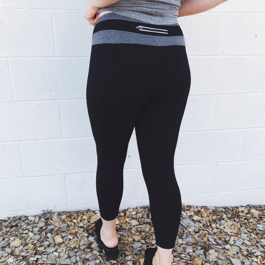 A person wears Wrap Band Leggings • 1070—black capris with a distinctive gray waistband and a back zipper pocket—while facing a white brick wall on mulch-covered ground.