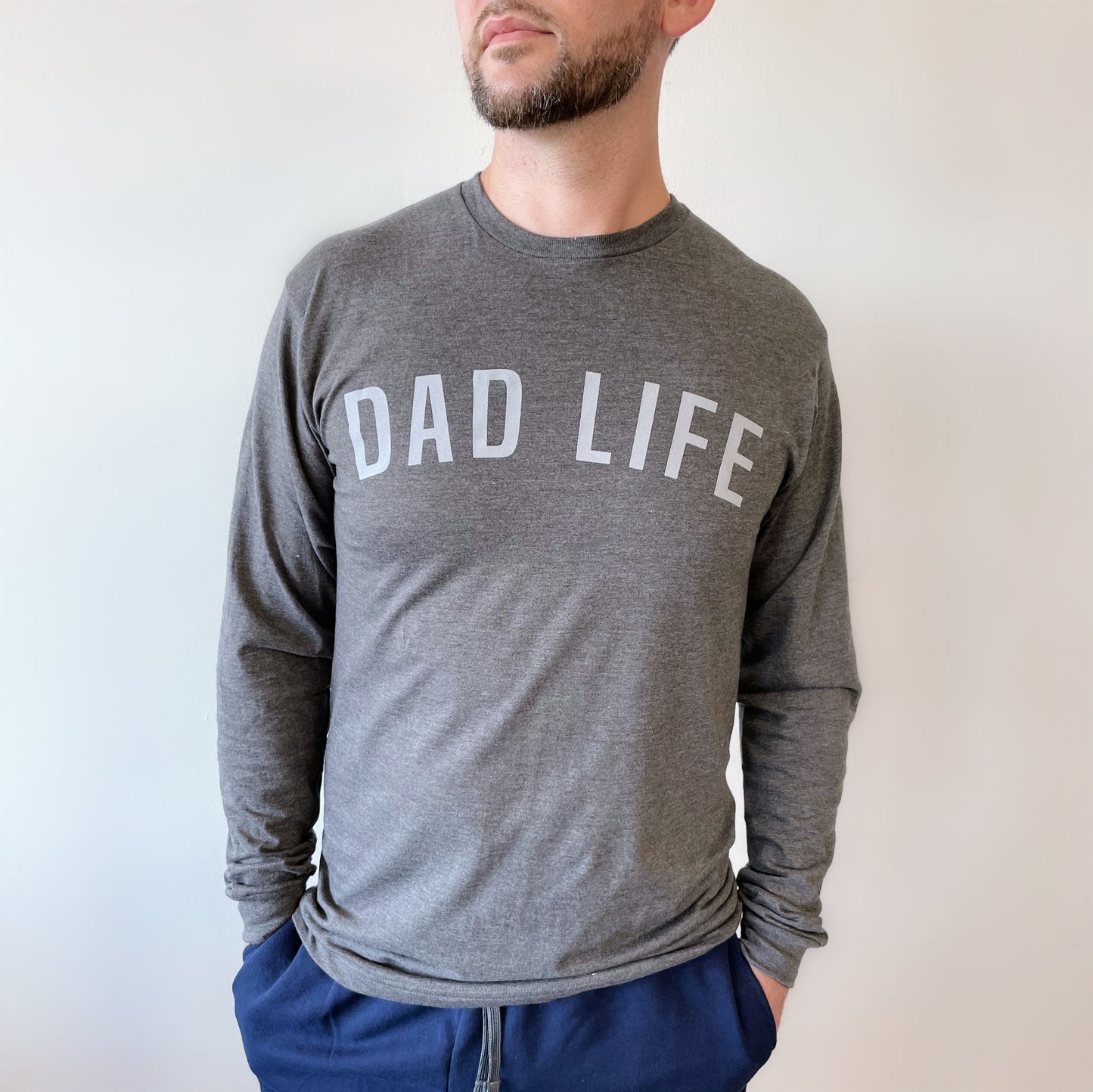 A man stands against a white background with his hands in his pockets, wearing the Dad Life Block • Charcoal Long Sleeve—crafted from soft ring-spun cotton and featuring DAD LIFE in bold white letters on the front.