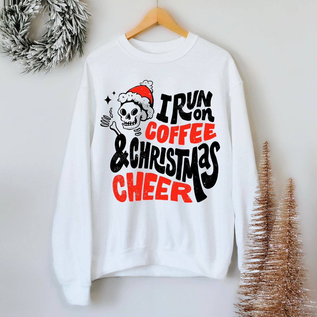The Christmas Cheer • White Pullover is a unisex white sweatshirt featuring a skeleton in a Santa hat with coffee, surrounded by wreath and gold trees. I run on coffee & Christmas cheer appears in black and red festive lettering.