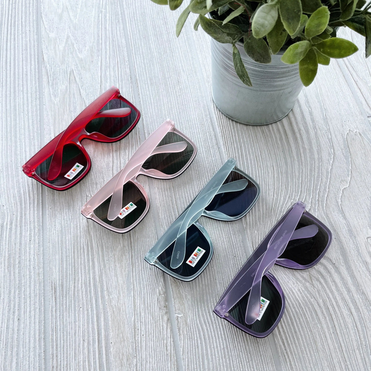 Four pairs of Arden Mirrored Sunnies • Kids, featuring UV protection and red, pink, gray, and purple frames, are arranged diagonally on a light wooden surface beside a green potted plant.