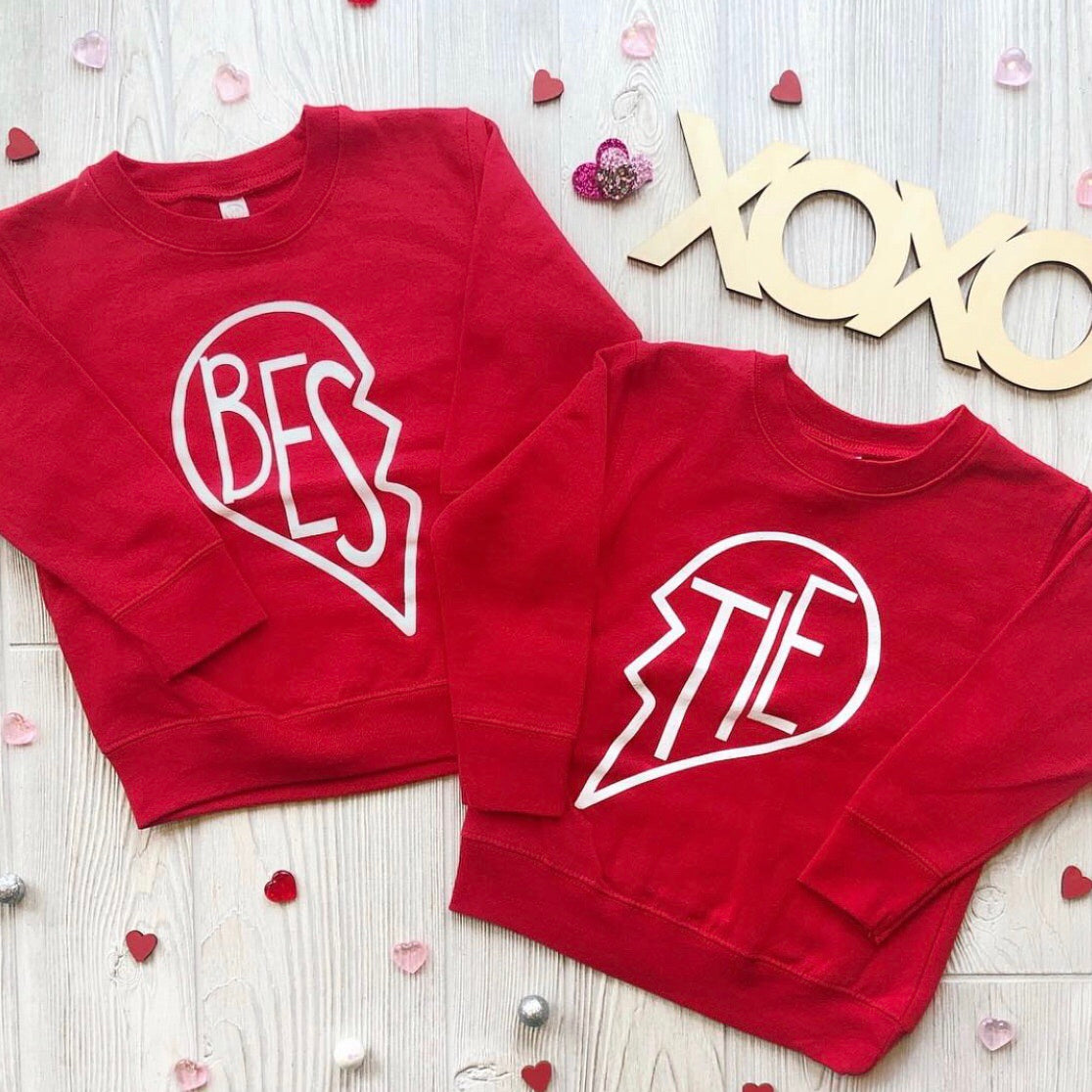 Two red Bestie Pullover • Red youth pullovers, unisex fit, lay side by side to form BESTIE inside a broken heart outline—surrounded by hearts and a wooden XOXO sign, perfect for celebrating friendship.