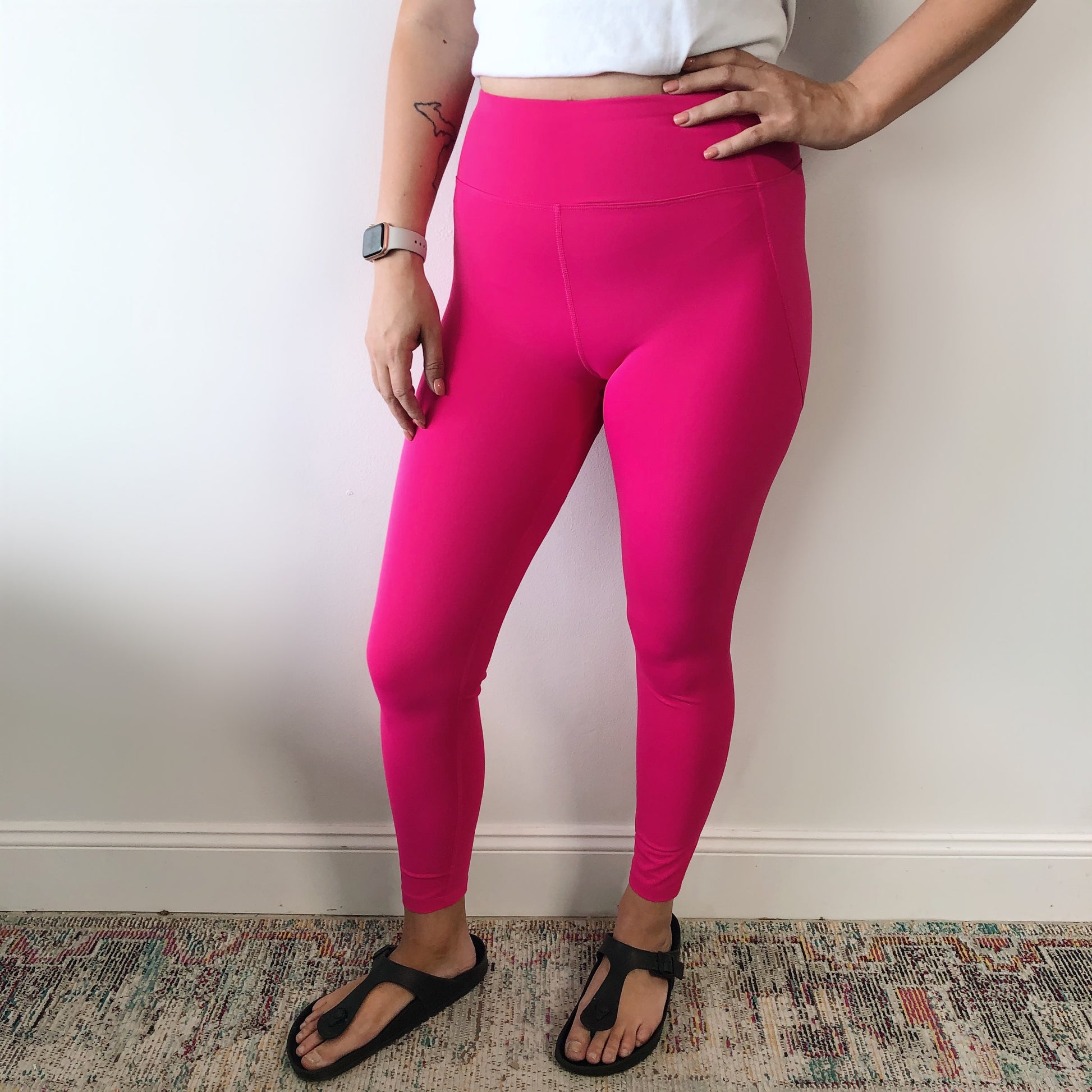 Indoors, a person poses with one hand on their hip, showing a small forearm tattoo. They wear bright pink Billie Legging 6209, a white cropped top, black sandals, and a watch. The background features a plain wall and patterned rug.