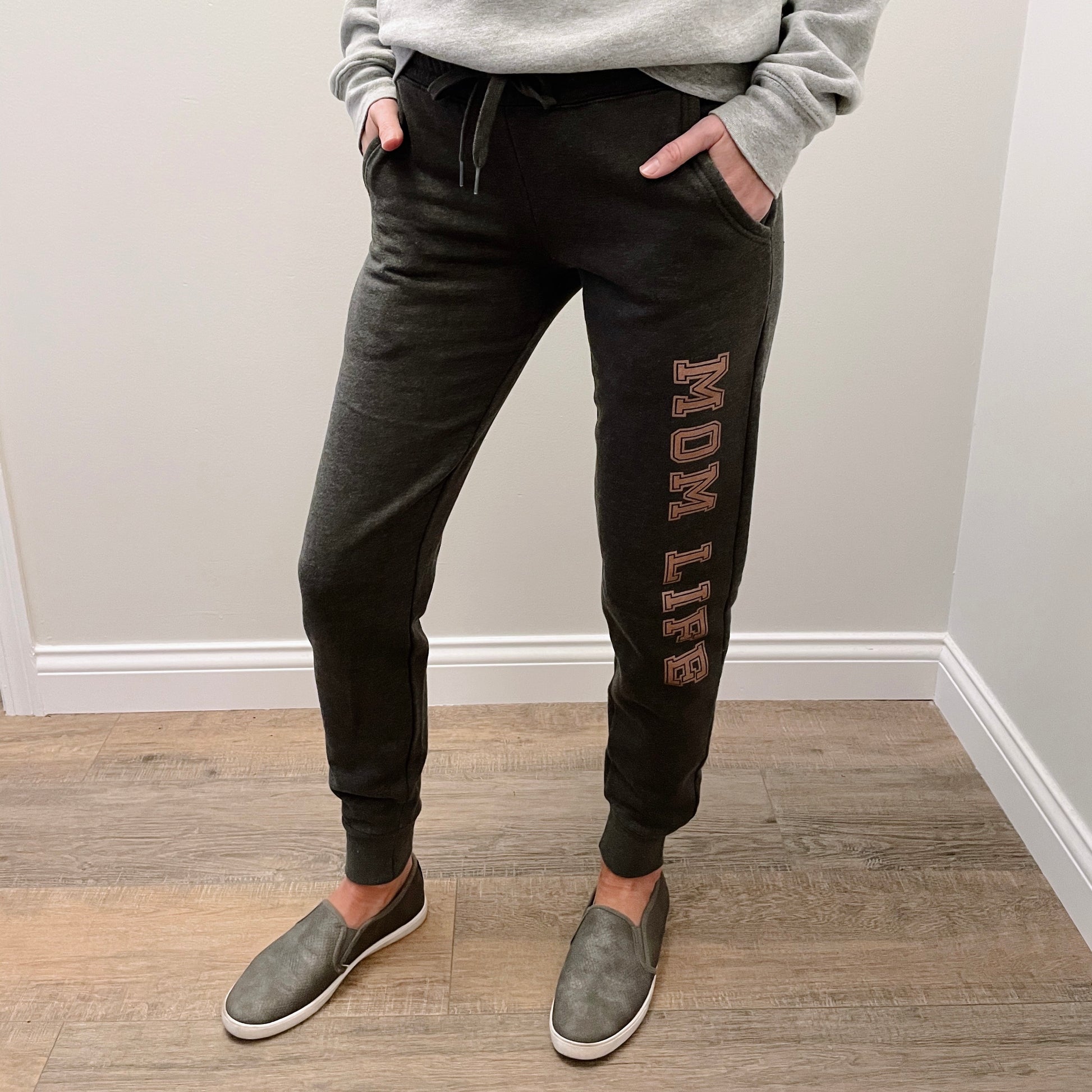 A person stands on a wooden floor wearing the MOM LIFE • Charcoal Jogger, a gray sweatshirt, and gray slip-on shoes. Hands are in pockets, showing only the lower half of the body.
