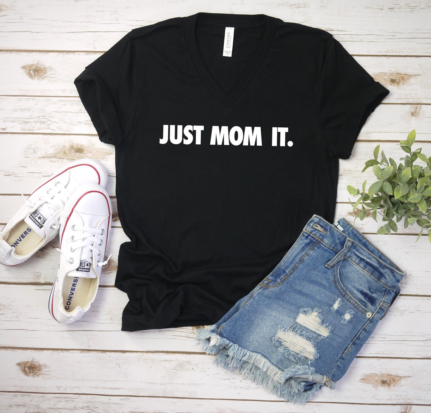The Just Mom It • Black V-Neck, featuring white lettering, is displayed with white Converse, ripped denim shorts, and a green plant on a white wooden surface—a must-have comfy unisex mom tee.