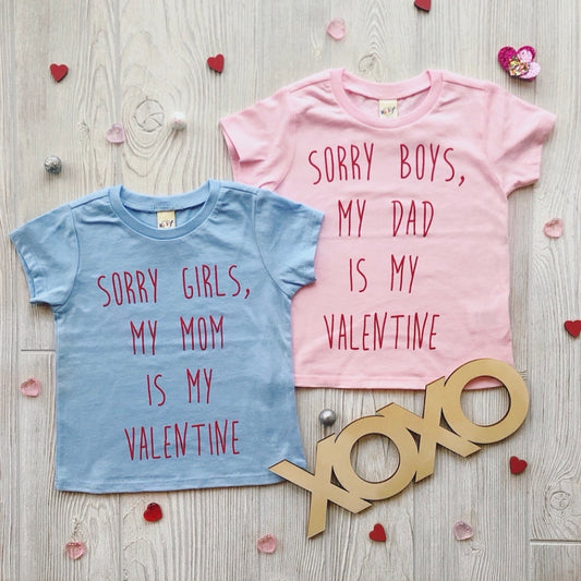 Two Sorry Tee • Blue or Pink kids shirts on a wooden floor with heart confetti. The blue tee reads Sorry girls, my mom is my valentine, and the pink says Sorry boys, my dad is my valentine. Wooden XOXO decor included.