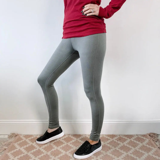 A person stands indoors on a patterned rug, wearing a long-sleeve red shirt and the Basic Sage Leggings • 6168 with a yoga style waistband, paired with black slip-on shoes. The photo is cropped from the shoulders down.