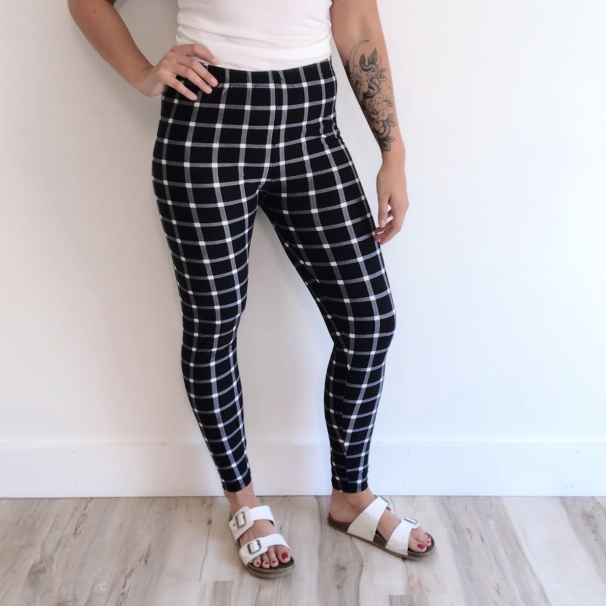 A person stands against a white wall wearing Grid Leggings with an elastic waistband, a white top, and white sandals. A tattoo is visible on their left arm. Only the lower half of their body is shown.