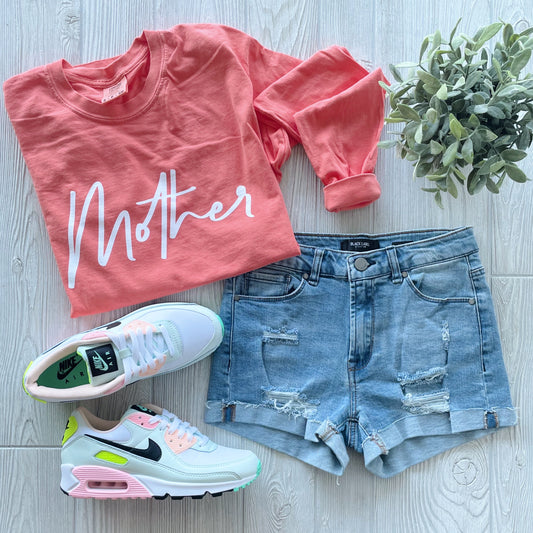 The Mother • Coral Long Sleeve shirt with white script, styled with distressed denim shorts, pastel-accented white Nike sneakers, and a small green potted plant on a light wooden surface.