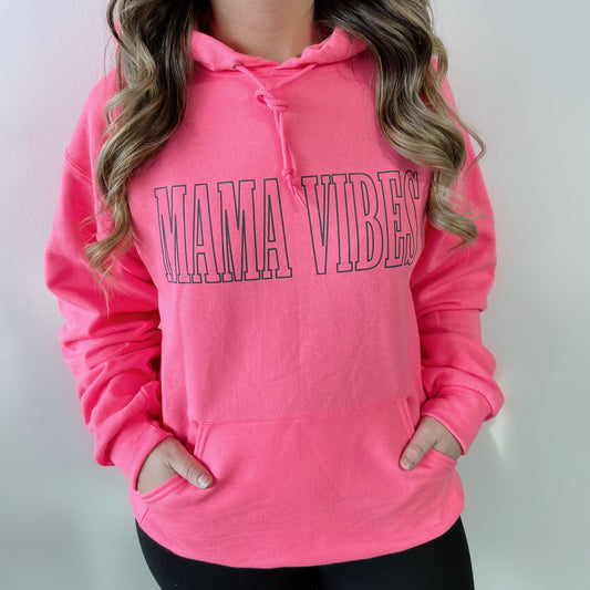 Someone with wavy brown hair wears the MAMA VIBES • Hot Pink Hoodie, a unisex neon pink sweatshirt featuring bold black MAMA VIBES lettering, with their hands tucked into the front pocket.