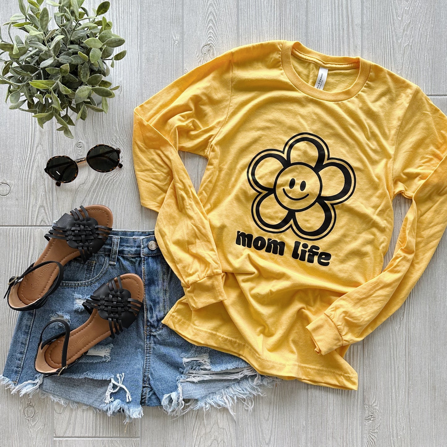 The Happy Flower • Yellow Long Sleeve features a cheerful flower and mom life text, styled with ripped denim shorts, black sandals and sunglasses, plus a potted plant on a light wood surface for a bright, playful vibe.