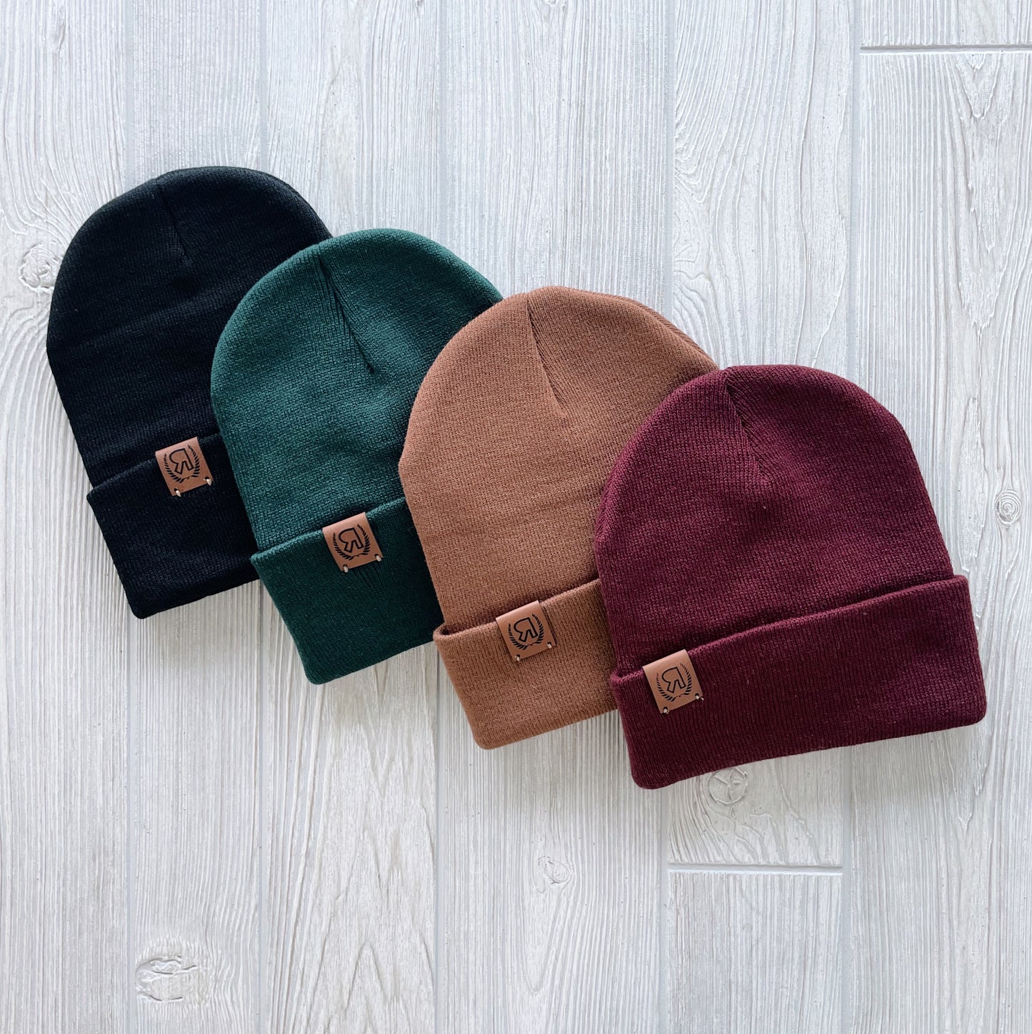Four Explore Collection Beanies in black, dark green, brown, and burgundy lie side by side on light wood. Each hand-sewn beanie has a small leather logo patch on its folded brim.