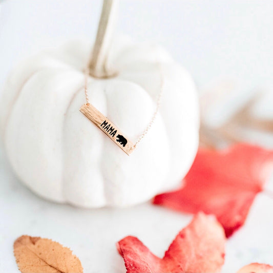 The Mama Bear • Bar Necklace features a rectangular pendant engraved with MAMA and a bear silhouette. Available in silver or gold, it includes an adjustable clasp for the perfect fit. Styled here on a white pumpkin among autumn leaves.