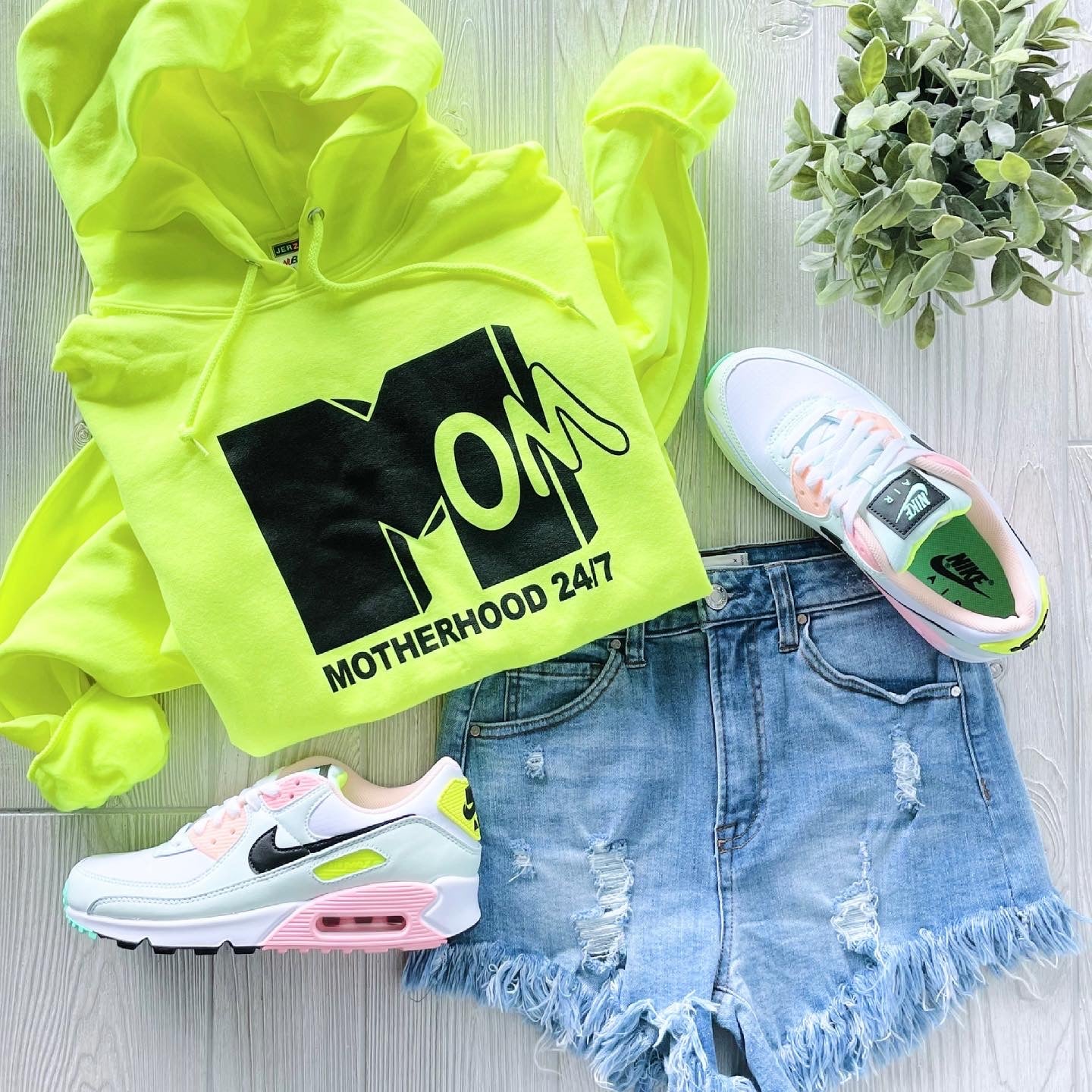 The Mom 24/7 • Neon Hoodie, distressed denim shorts, pastel-accented white Nike Air Max sneakers, and a small potted plant are displayed on a light wood surface.