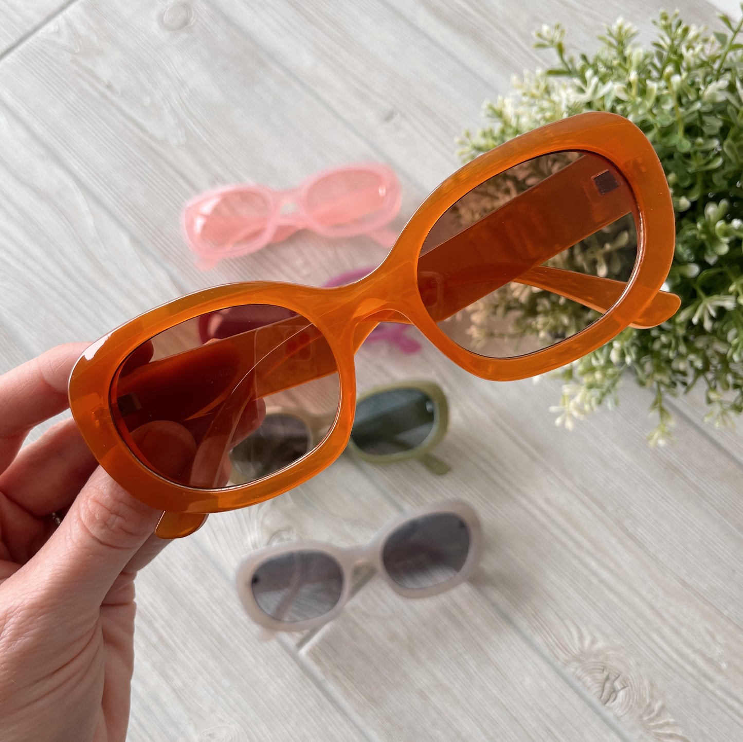 A hand holds orange Poppy Sunglasses • Adult • More Colors! above a light wooden surface, with four other colorful pairs—pink, green, gray, and beige—in the background near a small green plant. Final Sale Item.