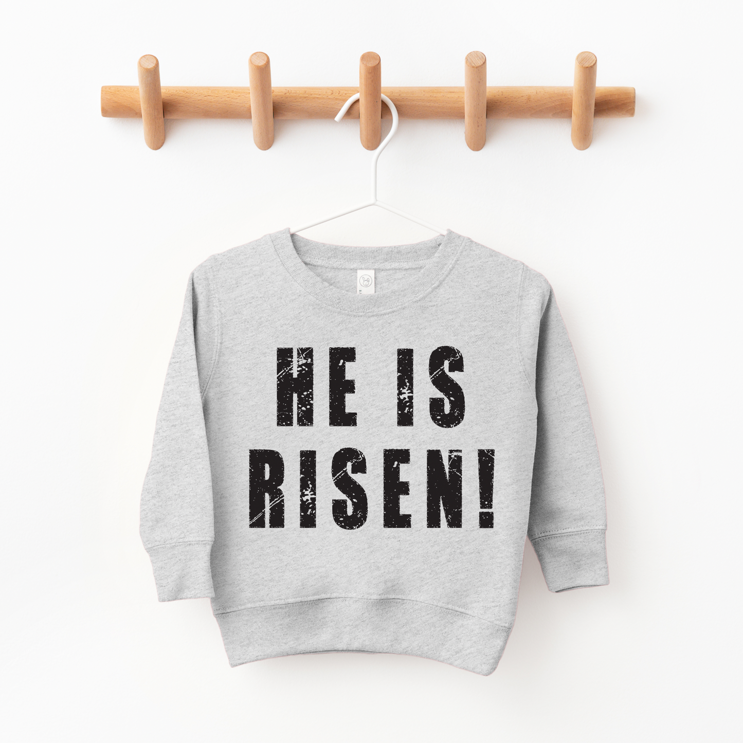 The He is Risen! Pullover, a gray unisex children’s sweatshirt with bold black text, hangs on a wooden peg rack—perfect for meaningful shopping and supporting small businesses.