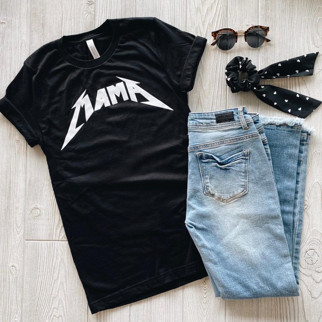 The Metal Mama • Tee, a black unisex top with “MAMA” in a bold metallic-style font, is styled with light blue jeans, sunglasses, and a black polka dot scrunchie scarf on a light wooden floor.