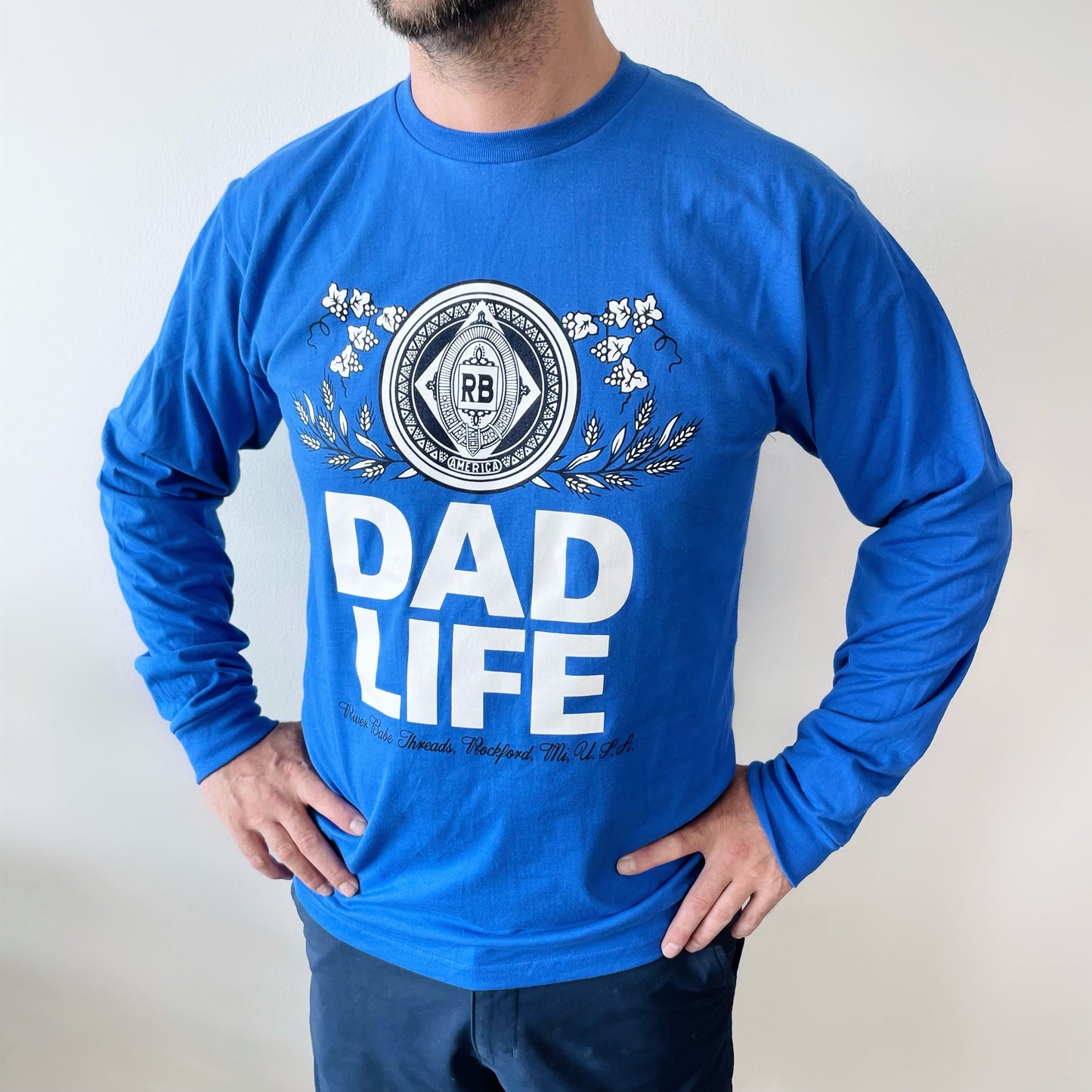 A man wears the DAD LIFE Royal • Long Sleeve, a bright blue classic crew neck shirt featuring bold white DAD LIFE text and a decorative circular crest, made in Michigan, standing against a plain white background.