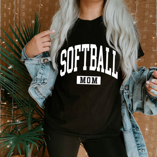 A person with long, wavy, light hair wears the Softball Mom • Tee from the Sports Mom Collection and a denim jacket. The backdrop includes a leafy plant and a tan woven screen decorated with string lights.