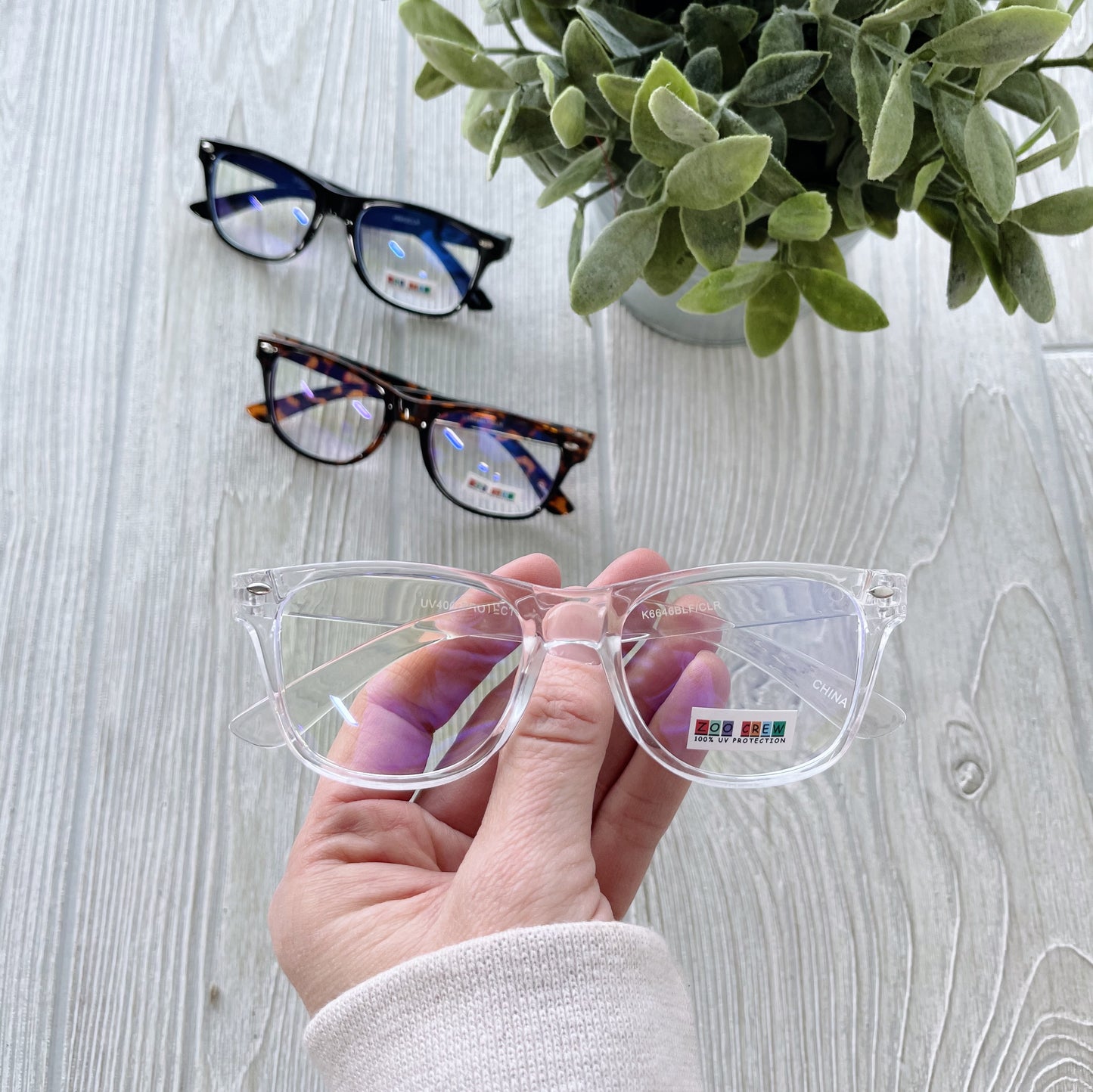 A hand holds a pair of Kids Classic • Blue Light Glasses above a light wooden surface, while two more pairs—one black and one tortoiseshell with UV protection—rest on the table beside a potted green plant.