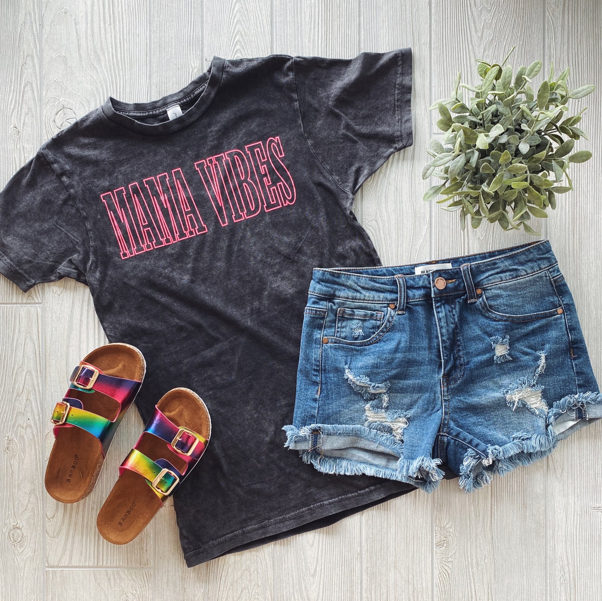 A flat lay featuring the black MAMA VIBES • Rocker Tee, distressed denim shorts, colorful metallic-strap sandals, and a small potted plant on a light wooden floor.