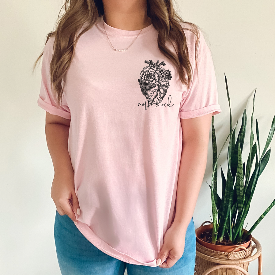 A person wearing the Heart Motherhood • Pink Tee, made from 100% Airlume cotton and featuring a floral graphic and “motherhood,” stands by a green potted plant, holding the hem against a plain background.