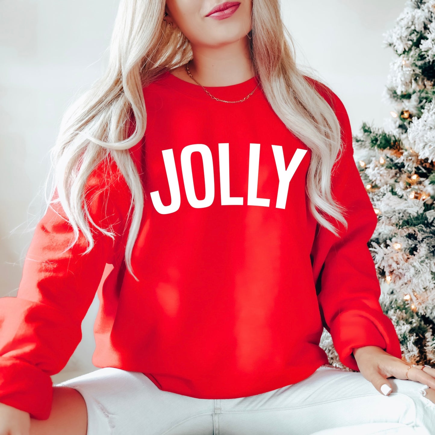 A woman with long blonde hair wears the Jolly • Adult Pullover in bright red, sitting near a decorated Christmas tree. Her face is partially out of frame.