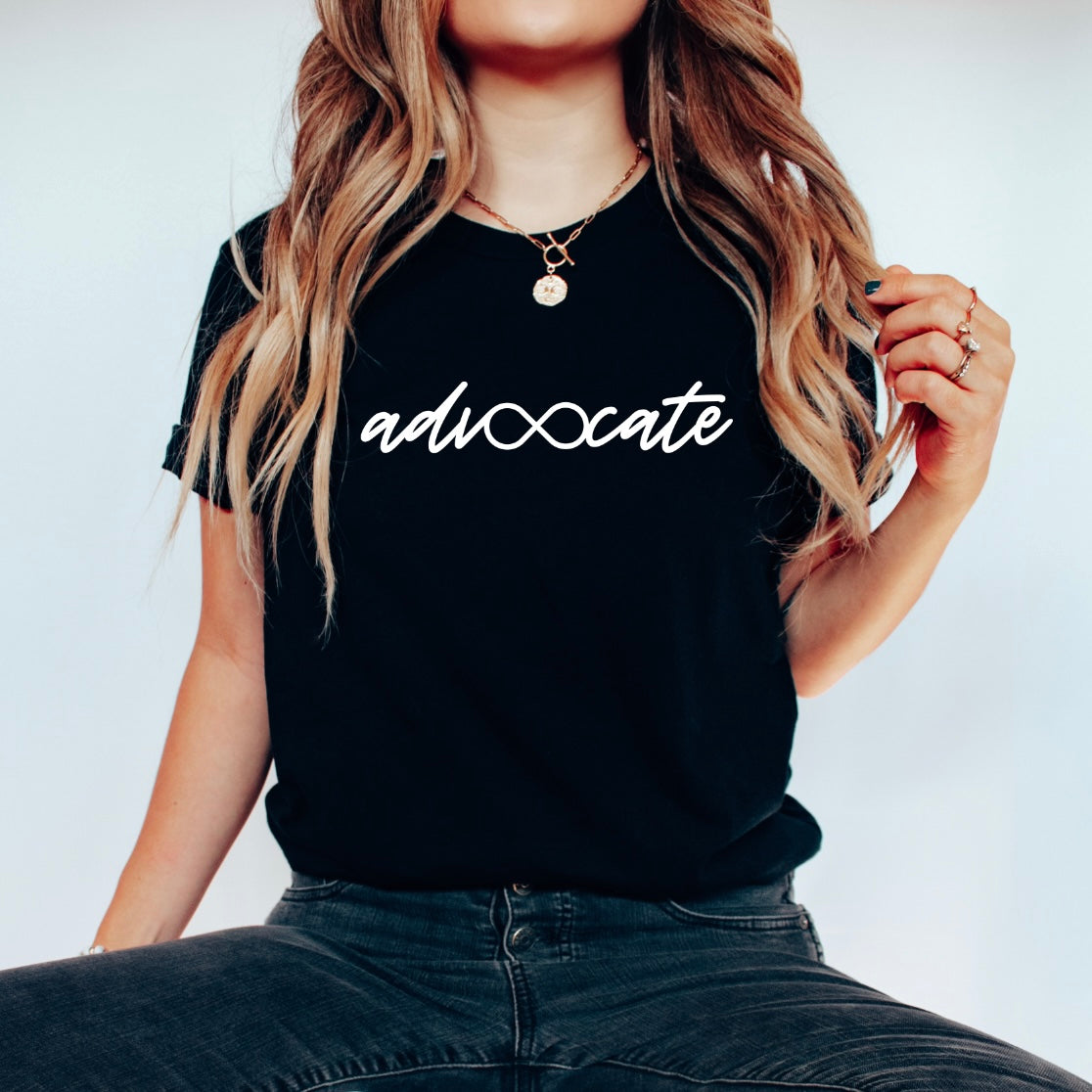 A woman with long wavy hair sits cross-legged wearing the Advocate • Black Tee, which features adv∞cate with an infinity symbol. She accessorizes with layered necklaces and rings.