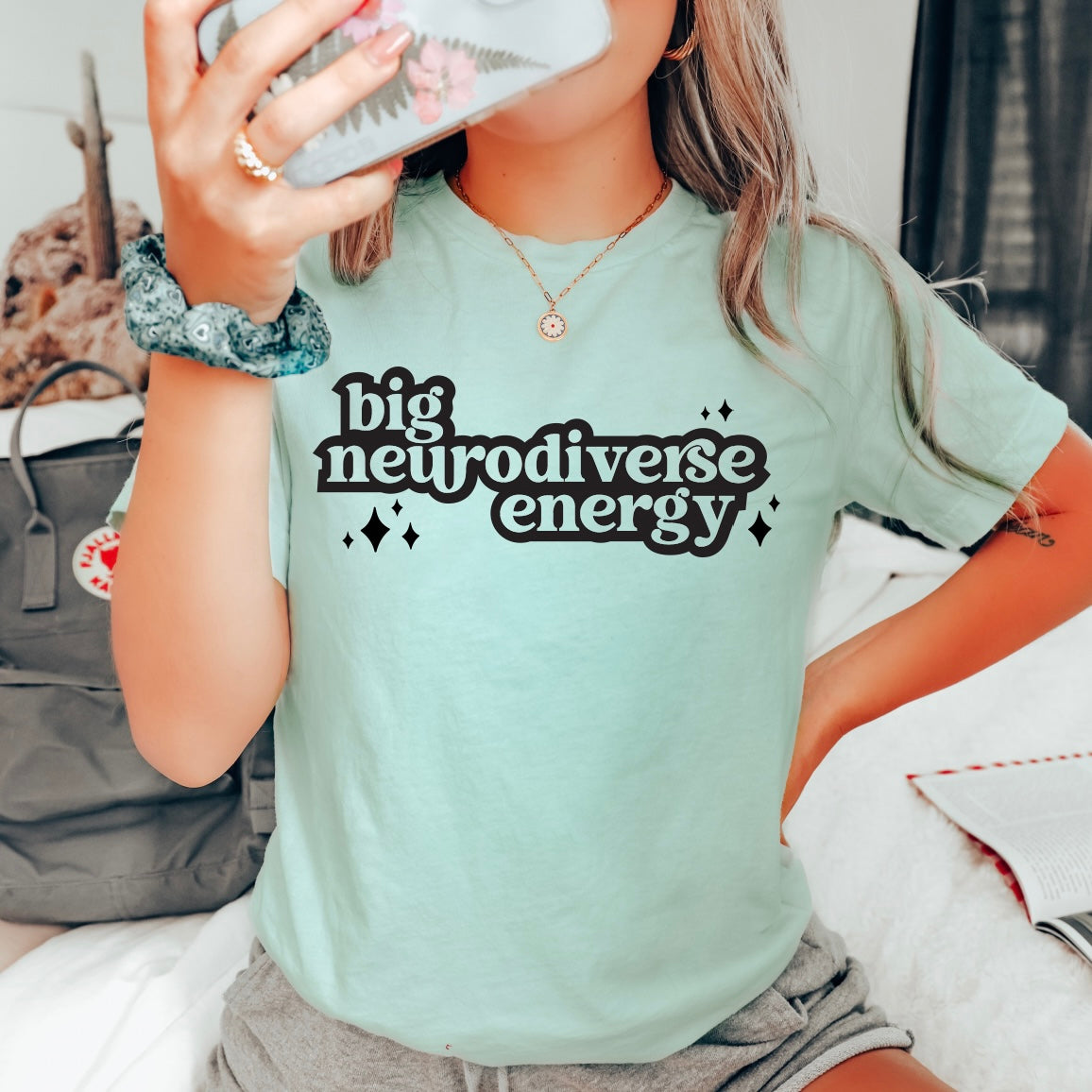 A person wearing a dusty blue Neurodiverse Energy tee takes a mirror selfie. The unisex retail fit shirt pairs perfectly with a scrunchie and gray backpack seen in the background.