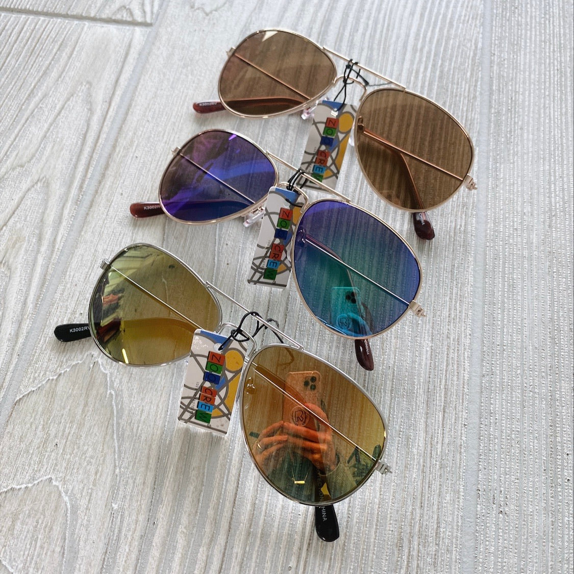 Three pairs of Aviator Sunnies • Kids with UV protection—featuring brown, blue, and yellow lenses—are displayed on a light wood surface. Tags attached; photographer’s reflection visible in the lenses. Final sale—don’t miss out!.