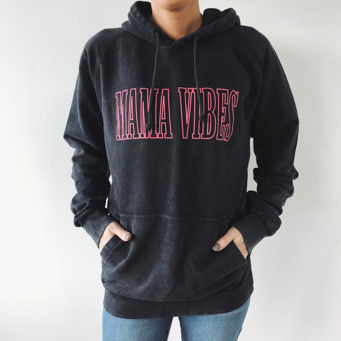 Wearing the MAMA VIBES • Rocker Hoodie, a vintage washed black sweatshirt with bold pink MAMA VIBES lettering, this person stands against a plain white background with hands in the pocket.