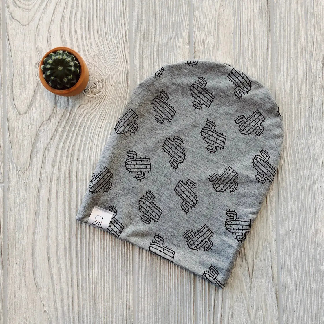 The Cactus Beanie, a handmade gray hat with a black cactus pattern and small white tag, rests on light wood beside a potted cactus—perfect for any River Babe who loves cozy style.
