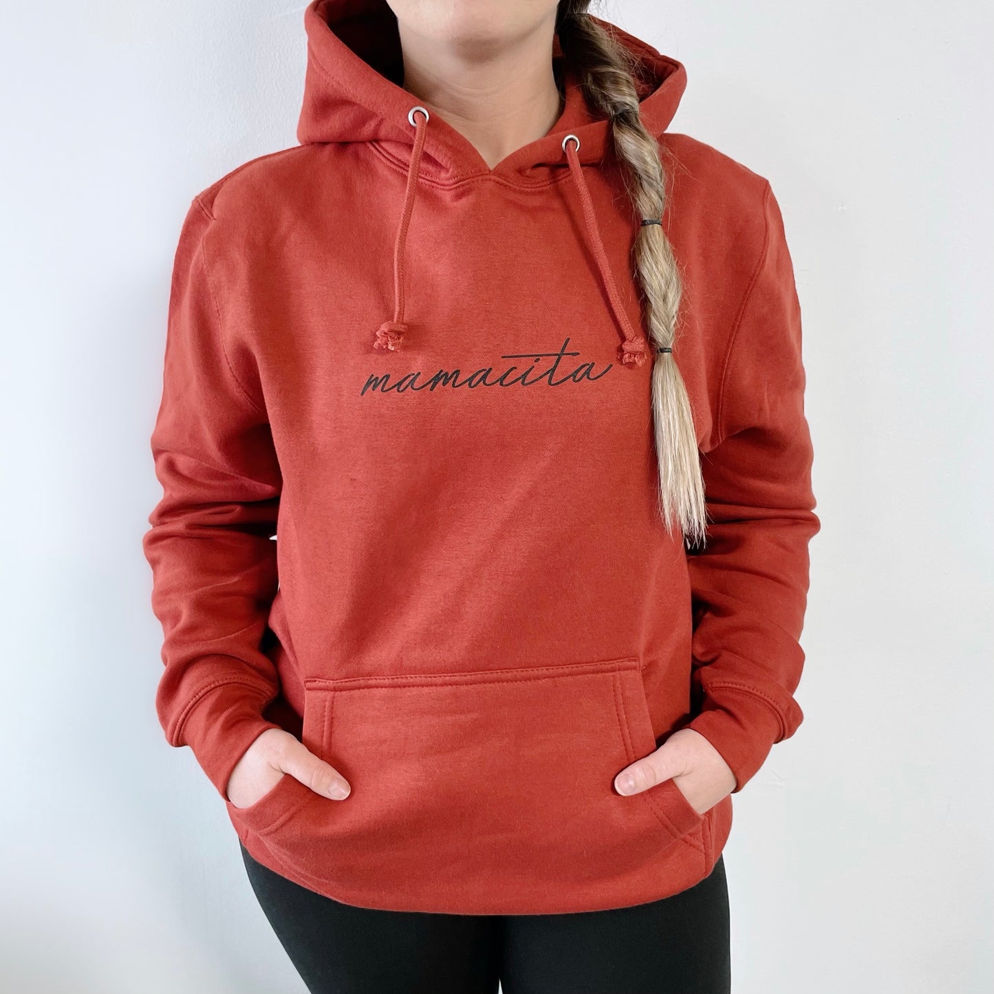 A person in a cozy, unisex Mamacita • Terracotta Hoodie with script print on the front stands against a white background. The red hoodie has a front pocket, and the wearer’s long blonde braid is visible as they tuck their hands in the pocket.