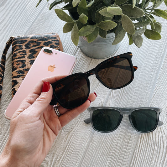 A hand with red nail polish holds Simon Sunglasses • Adult • More Colors! with UV400 Protection above a table featuring a pink iPhone, leopard print wallet, gray sunglasses, and a potted plant.