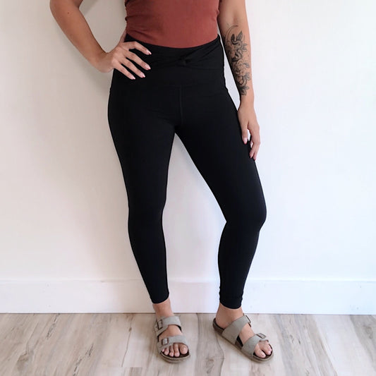 A person stands indoors against a white wall wearing a rust top, the Twist Accent Highwaist Legging • 2869 in black with tummy control, and light brown sandals. One arm is bent at the waist, revealing a forearm tattoo. The floor is light wood.