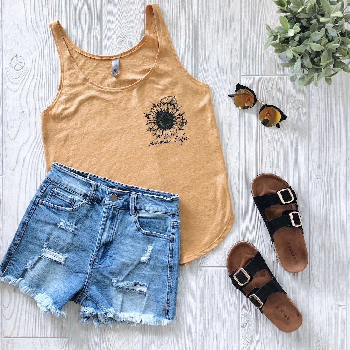 Flat lay of a summer outfit: Sunflower • Gold Tank with Mama Life Sunflower text, flowy yellow fit, paired with distressed denim shorts, black buckle sandals, sunglasses, and a green plant on a light wooden floor.