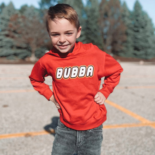 A young boy smiles outdoors in the Bubba • Lego Hoodie—a bright red hoodie featuring bold white BUBBA lettering. He stands with hands on hips in a parking lot with trees in the background.