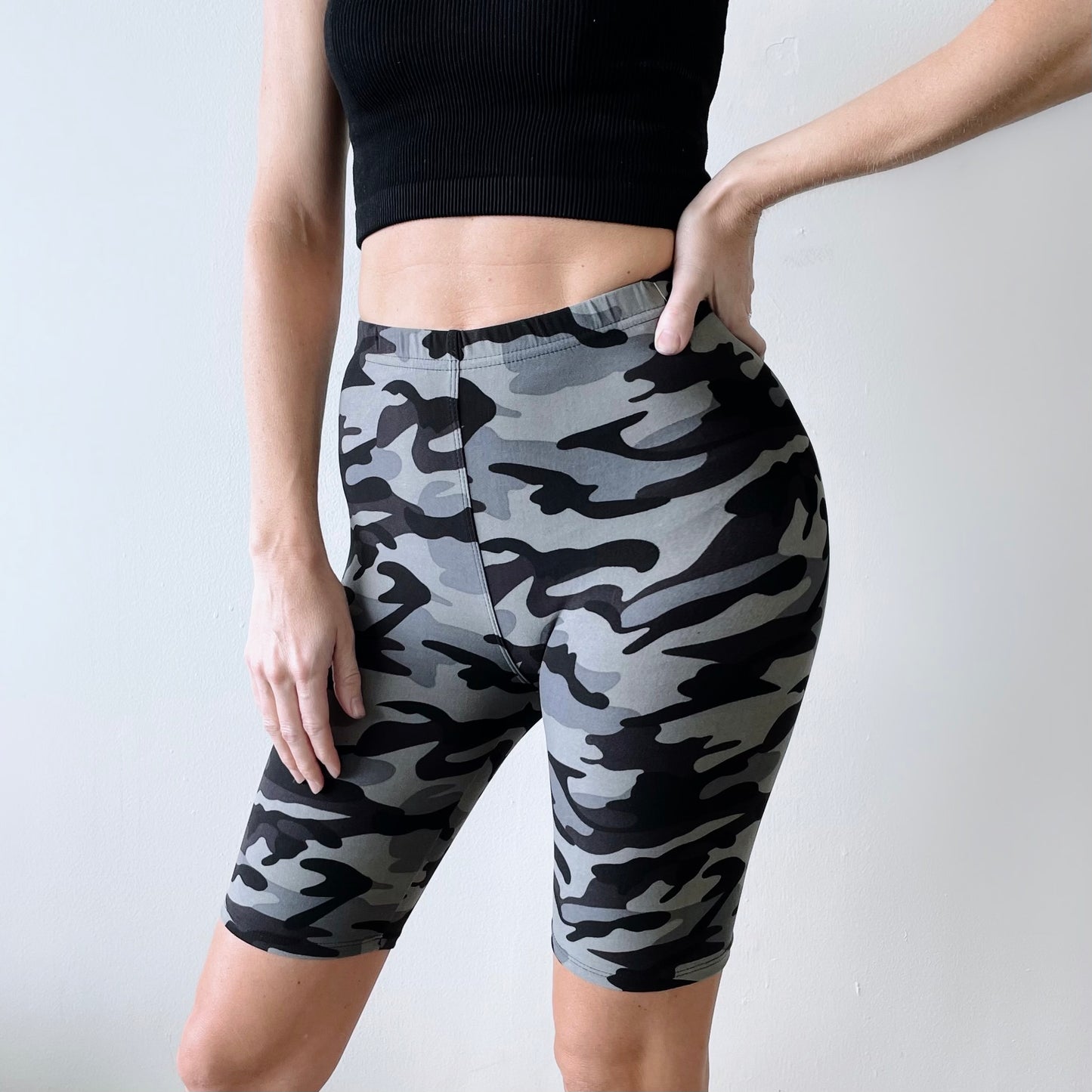 A person wearing a black crop top and Light Gray Camo • Brushed Biker Shorts stands against a plain white wall with one hand on their hip.