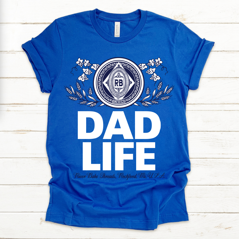 The DAD LIFE • Royal King Tee is a blue crew neck made from soft ring-spun cotton, unisex sizing, with a decorative circular emblem, floral accents, and bold white DAD LIFE text. River Babe Threads, Rockford, TN USA appears in smaller print.