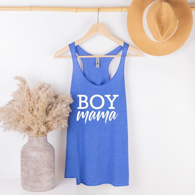 The New Boy Mama • Royal Tank, a blue racerback top with BOY Mama in white, hangs on a wooden hanger beside a straw hat and dried pampas grass—ideal for casual women’s style.