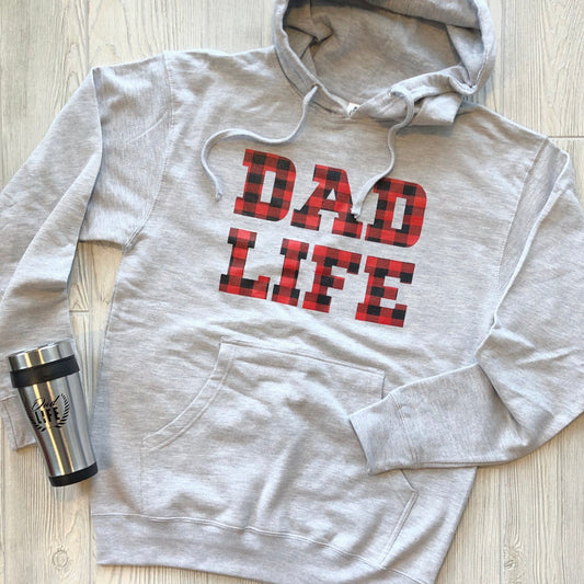 The Dad Life • Plaid Hoodie in ash gray features a bold red and black plaid DAD LIFE design and rests beside a sleek silver travel mug—perfect for elevating your fall wardrobe.