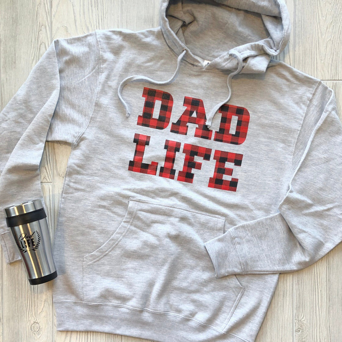 The Dad Life • Plaid Hoodie in ash gray features a bold red and black plaid DAD LIFE design and rests beside a sleek silver travel mug—perfect for elevating your fall wardrobe.