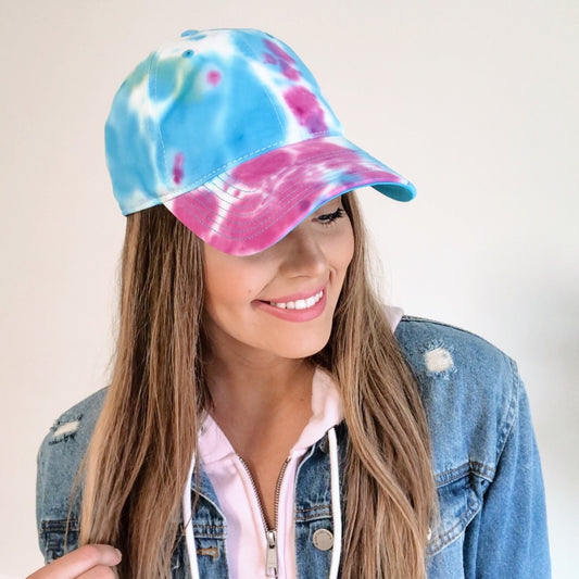 A woman smiles and looks down against a plain white background, wearing the Tie Dye Hat • Jelly Bean, a light pink hoodie, and a denim jacket.