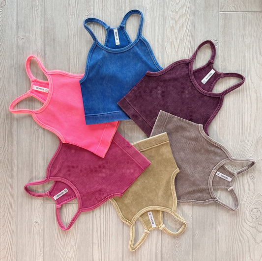 Six Luna Vintage Crop tops from the Aurora Collection in blue, burgundy, brown, olive, pink, and mauve are arranged in a circle on a light wooden floor, front side up to highlight their stretchy ribbed fabric.