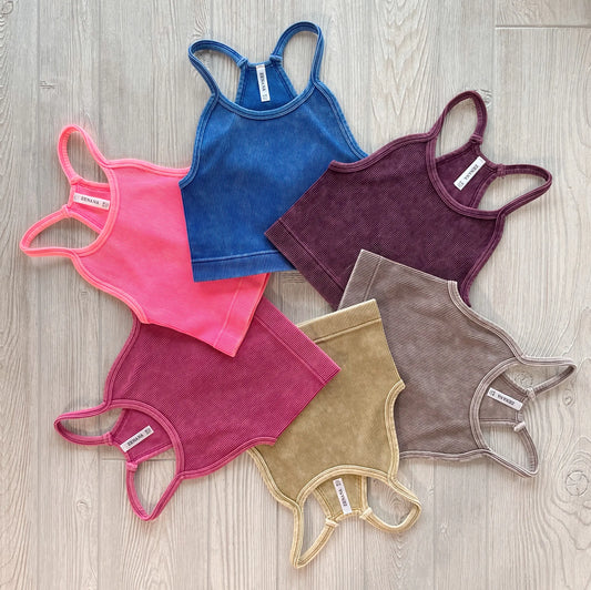 Six Luna Vintage Crop tops from the Aurora Collection in blue, burgundy, brown, olive, pink, and mauve are arranged in a circle on a light wooden floor, front side up to highlight their stretchy ribbed fabric.