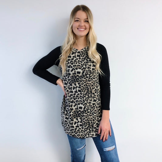 A woman with long blonde hair smiles against a white wall, wearing a Long Sleeve Cheetah Raglan (SIZE SMALL) with black sleeves and blue ripped jeans. The loose fit is highlighted as she rests one hand on her hip.