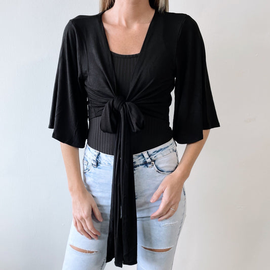 A woman models the Jasmine Cardigan in black, tied at the front over a ribbed tank and paired with light-wash ripped jeans. She stands against a plain light background, with her face not visible. Available in more colors!.