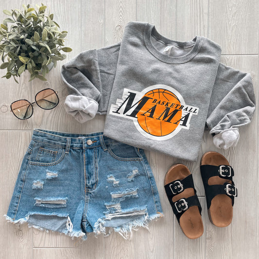 Flat lay of a casual outfit: Basketball Mama • Gray Pullover, distressed denim shorts, black buckle sandals, round glasses, and a small green potted plant on a light wood floor.