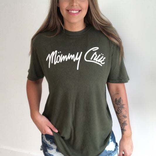 A woman with long light brown hair and a floral tattoo on her left forearm smiles while wearing the Mommy Crue • Vintage Olive Tee in a unisex style, paired with distressed denim shorts.