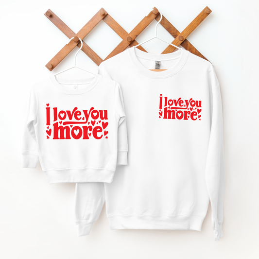 An adult-sized unisex “Love You More” white pullover hangs on a wooden hook, featuring red “I love you more” text with hearts on the front, shown against a white background.